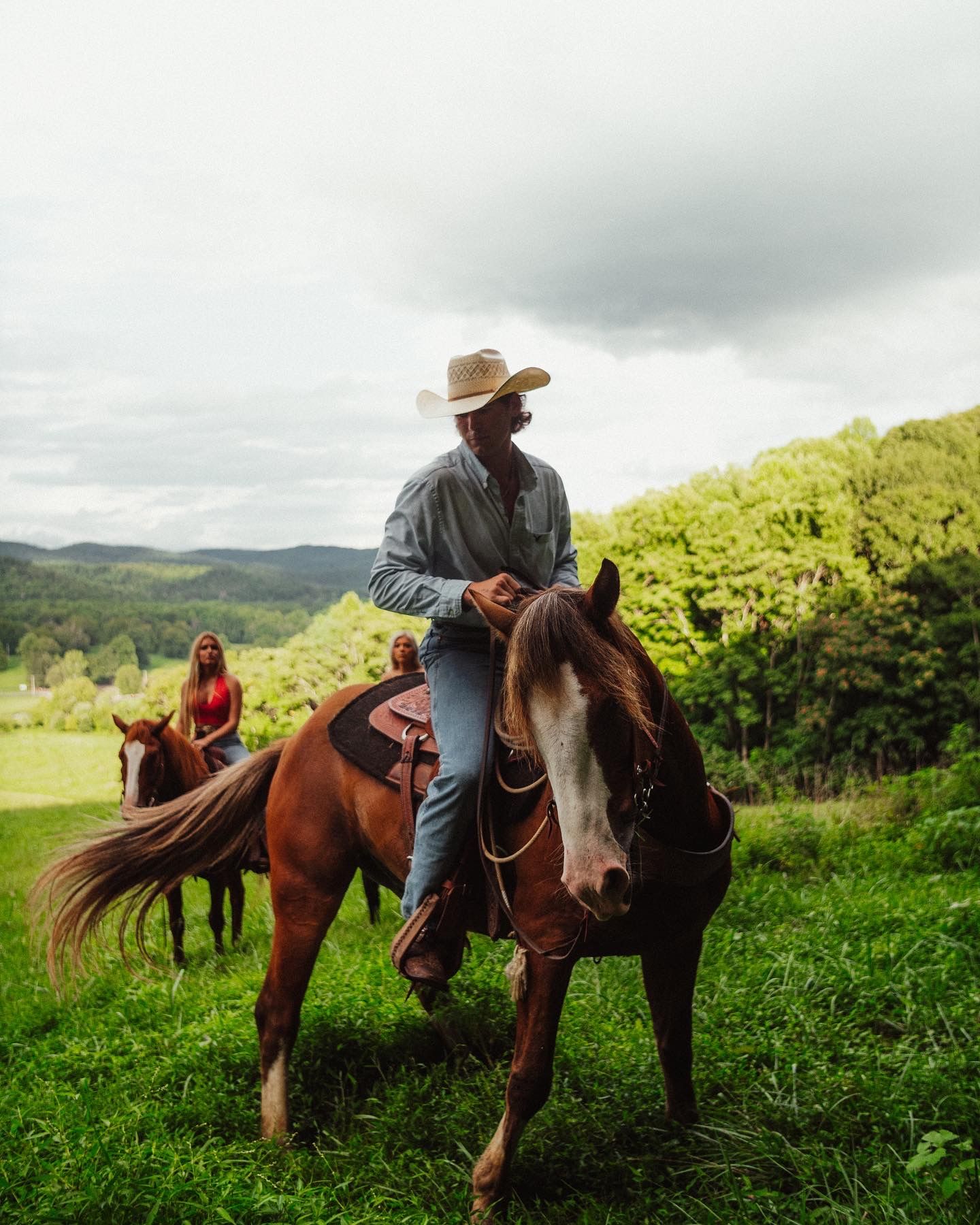 Jonah riding a sorrel horse through green hills in a straw hat, two riders behind him