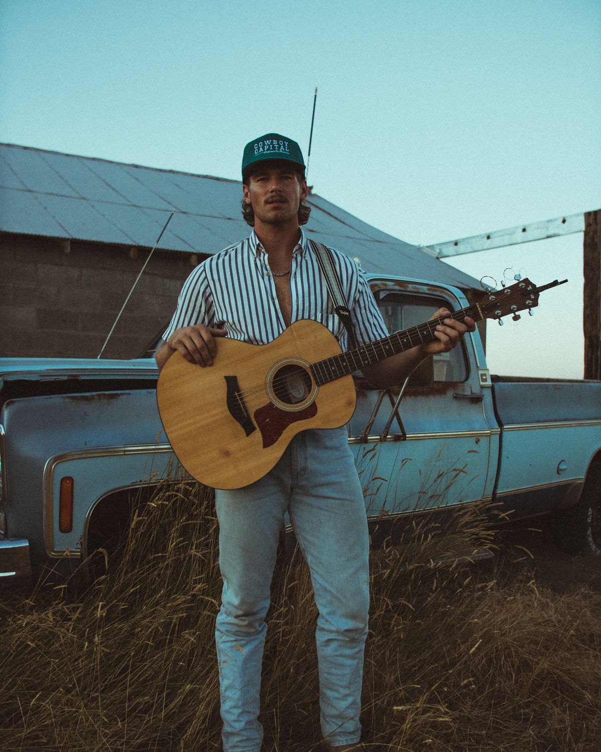 Jonah holding an acoustic guitar in front of an old blue pickup and a tin-roofed barn