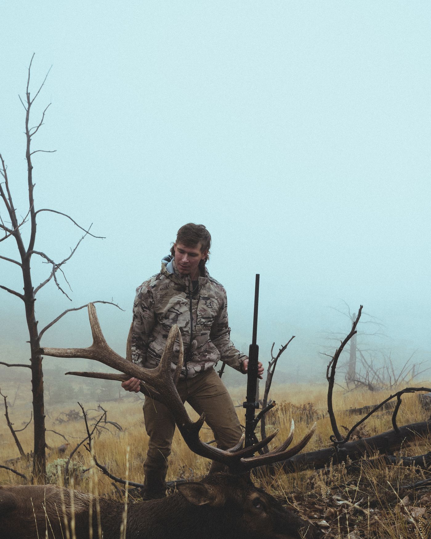 Jonah kneeling beside a bull elk in heavy fog among burned trees