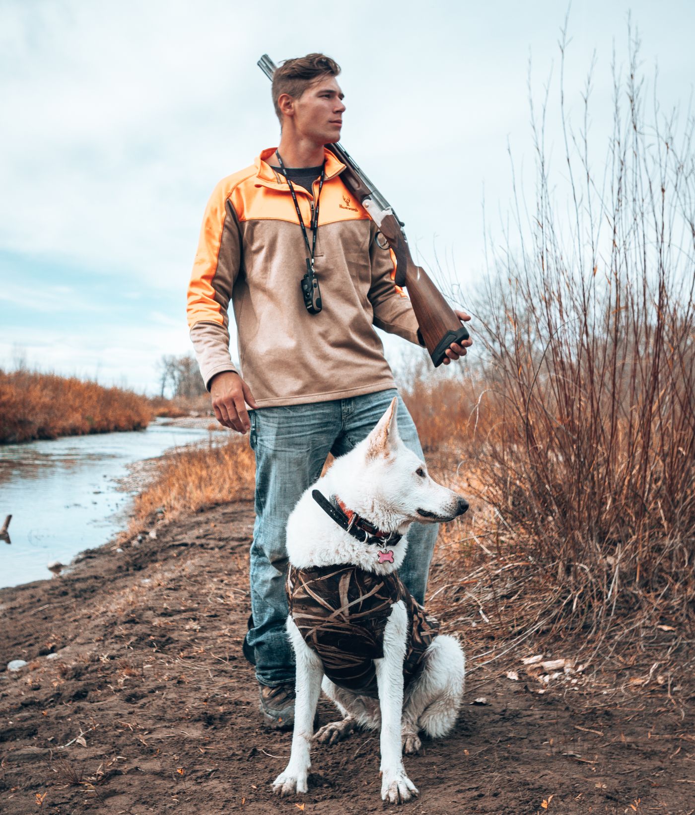 Jonah in a blaze-orange pullover with a shotgun on his shoulder, a white German Shepherd sitting at his feet by a river