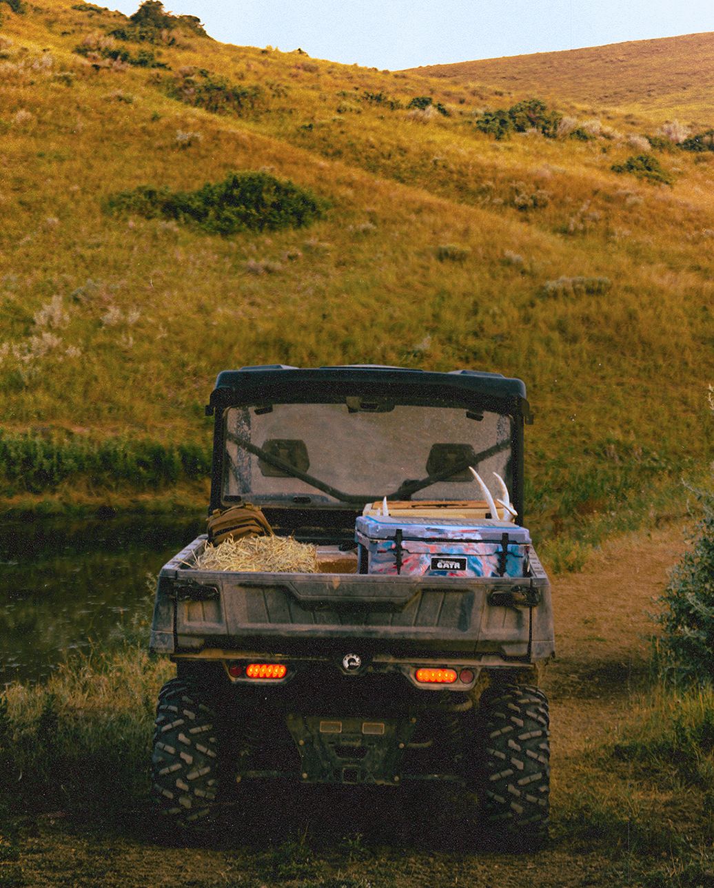 A muddy ranch UTV with hay, a cooler and antlers in the bed, parked by a pond below a grassy hill