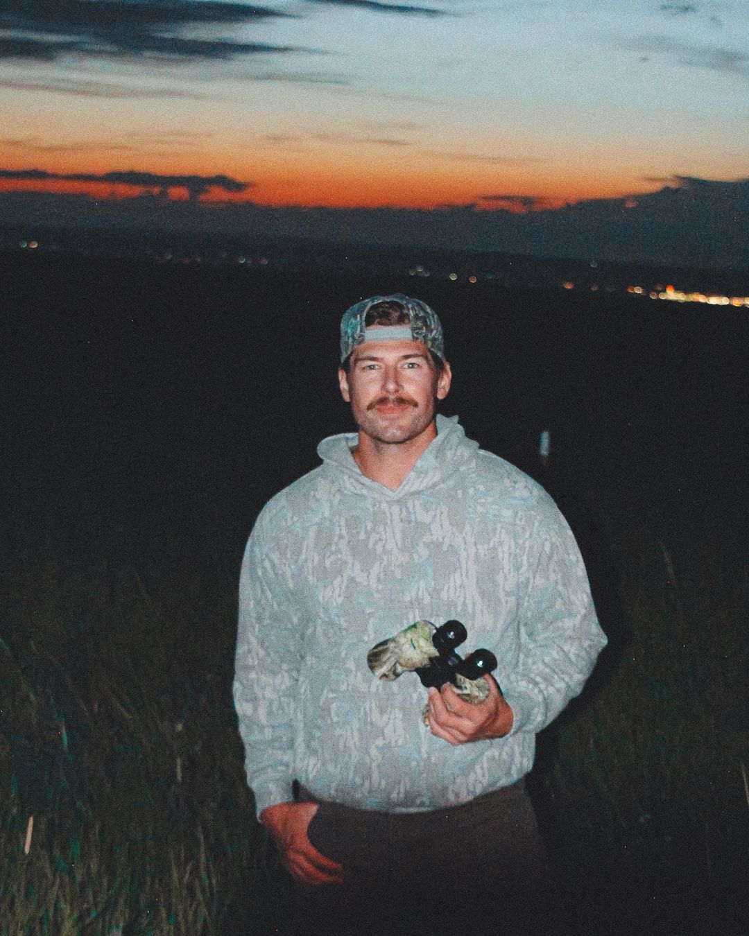 Jonah in a camo hoodie holding binoculars on a hillside at dusk, city lights far below